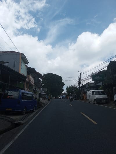 The image captures a daytime street scene in Ura-Dozhi, Bhutan, under a partly cloudy sky with prominent white cumulus clouds. An asphalt road, marked with a solid yellow centerline transitioning to a broken yellow line, and a solid white edge line, occupies the foreground and recedes into the distance.
On the left side of the road, a blue minivan is parked partially on the concrete curb next to a drainage gutter. Behind it, a row of two-story buildings, exhibiting varied architectural styles and painted facades, including one with light blue and orange sections, feature balconies and numerous overhead utility wires. A large, dense-foliage tree is visible further down the street on this side.
On the right side of the road, a white panel delivery van is parked. Further along this side, a building features a prominent "J&T CARGO" sign. Additional structures, obscured by a dense network of overhead wires, line the street, along with a street light pole.
One individual, wearing a helmet, is operating a black motorcycle on the road, moving away from the camera in the right lane. Several other vehicles, including cars, are visible in the distant background, also moving away. Indistinct figures are observable near the buildings lining the street in the distance.