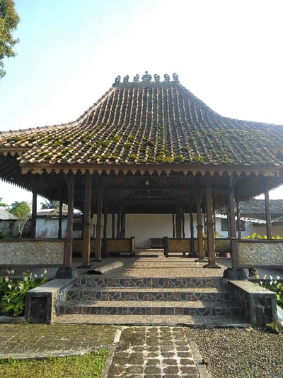 An outdoor pavilion, located in Dulce Nombre de Jesús, Costa Rica, is depicted in a full frontal, eye-level view under a clear, bright sky. The structure features a prominent multi-tiered, brown tiled roof, extensively covered in patches of green moss, with a series of eight ornamental finials along the central ridge. Intricately carved or cut wooden eaves are visible beneath the roofline. The roof is supported by numerous dark-stained wooden posts.
The pavilion is elevated on a concrete platform, accessed by a central set of four steps surfaced with brown and grey mosaic-patterned tiles, flanked by low, dark-colored concrete or stone retaining walls. A low, textured, light-colored wall or railing encloses the perimeter of the elevated platform. Inside the pavilion, a plain white wall is visible at the rear, with two low, horizontal benches or platforms, possibly woven or bamboo, situated on a paved floor. No individuals are present in the scene.
In the immediate foreground, a rectangular paved pathway, partially covered in moss and grime, leads towards the steps. To the right of this pathway is an area of small gravel, and to the left is an area of mowed grass. Green foliage, including bushes and other plants, is visible on either side of the steps and around the base of the pavilion. A tree trunk and green leaves are visible in the upper left corner of the frame. In the background, beyond the pavilion, other residential-style buildings with light-colored walls and corrugated roofs are partially visible, alongside additional distant trees. The scene is illuminated by natural sunlight, casting shadows.