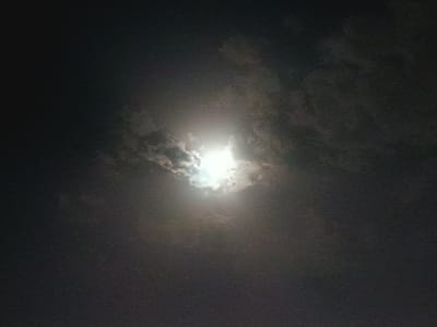 The image depicts a night sky with a bright, luminous moon partially obscured by dark clouds. The moon's light casts a glow that illuminates some of the surrounding cloud formations, creating a dramatic contrast between light and shadow. The overall scene suggests a cloudy night with a bright celestial body. The location is identified as Onaldy, Turkmenistan, but no specific landmarks or urban elements are visible in the image to support this. There are no people or other discernible subjects present.