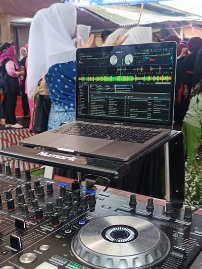 A forensic examination of the image reveals a DJ setup positioned outdoors or in a semi-enclosed public area in Kawungluwuk, Indonesia. The foreground displays the upper section of a black DJ mixer/controller, featuring numerous black rotary knobs, faders, a silver-colored jog wheel, and various illuminated buttons (some blue, some green). Directly above the mixer, a silver-gray MacBook Pro laptop is open and elevated on a black Numark-branded laptop stand. The laptop screen displays the Serato DJ Pro software interface, showing two virtual decks with colorful waveforms, BPM counters (117), track titles, and a music library.
In the mid-ground and background, multiple individuals are present. A woman wearing a white hijab and a blue patterned shirt is prominently positioned on the left, facing away from the camera. Other individuals, primarily women, some also wearing hijabs, are visible in varying states of focus and distance, suggesting a gathering or event.
The environment includes a blue and white banner with partially visible Indonesian text ("MENDUKUNG KEDAULATAN") in the upper background. Corrugated metal sheeting is also visible. The ground appears covered by a striped mat or rug on the left, while green foliage is visible on the far right, indicating an outdoor or semi-outdoor setting. No specific actions or interactions between individuals are discernible beyond general presence at an event.