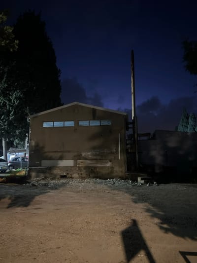 A tall, rectangular building with a pointed roof stands beside a dirt road at night. Several vertically stacked windows are visible on the building's facade. A long, thick pipe extends horizontally from the top of the building. Trees surround the building, adding a natural element to the urban setting. Parked cars can be seen near the base of the building. The sky is dark blue, suggesting nighttime.