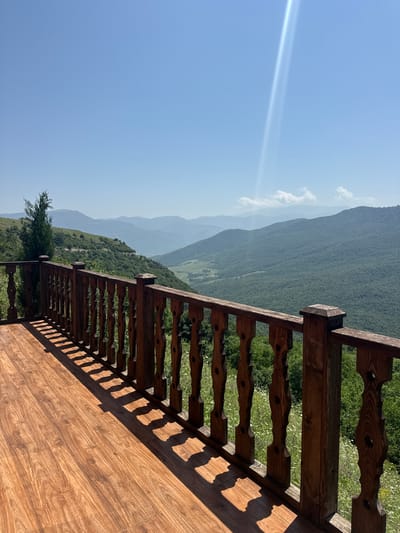 An outdoor perspective shot shows a wooden deck with a wooden railing on the left side. The railing is made of dark brown wood with ornate carvings on each spindle. The deck is made of polished wood planks with a light brown grain. Beyond the railing, a lush green valley stretches out, dotted with trees and wildflowers. In the distance, layers of mountains fade into a hazy blue, and the sky above is a clear, bright blue with a single, bright beam of sunlight shining from the upper right corner. The scene is likely captured from a vantage point in or near Yenokavan, Armenia, a location known for its natural beauty and mountainous landscape. There are no people or animals visible in the image.