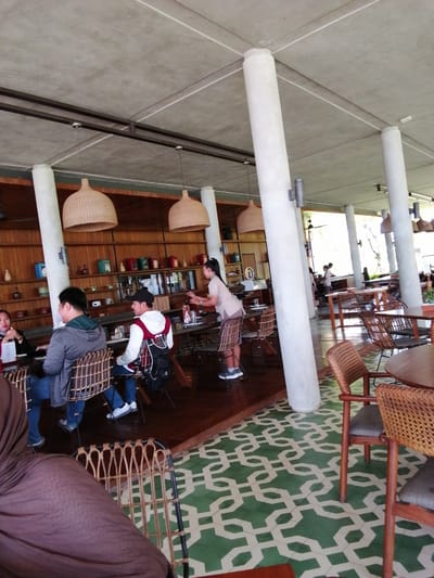 The image depicts the interior of a restaurant or cafe, likely located in Banyuwangi, Indonesia, characterized by its open-air design with a concrete ceiling and large support pillars. The flooring is a distinctive green and white patterned tile. Several large, woven pendant lights hang from the ceiling.
In the foreground, a person wearing a brown headscarf and covering is seated at a table, facing away from the camera. To the left, a man in a grey hoodie and blue jeans is seated at a table, looking towards the right. Next to him, another man in a white hoodie, a red vest, a black cap, and blue jeans is also seated at the table, facing the right. Both men are seated on rattan chairs.
In the mid-ground, a woman in a light-colored shirt and shorts is standing near a table, appearing to interact with something on the table. More tables and chairs are visible in the background, as well as wooden shelves filled with various decorative items. In the far background, other patrons and staff are present, and the environment appears to open to an outdoor area with vegetation visible. The overall atmosphere suggests a casual dining establishment with a natural or tropical aesthetic.