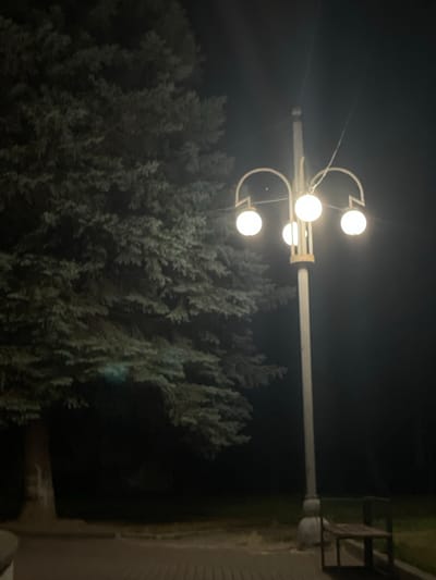 The image depicts a nighttime scene with a tall lamppost in the foreground, featuring four spherical lights. To the left of the lamppost is a large evergreen tree. In the background, a bench is visible. The ground appears to be paved. The lighting suggests an outdoor public space, possibly a park or a street in Stryi, Ukraine. The overall atmosphere is dark and quiet.