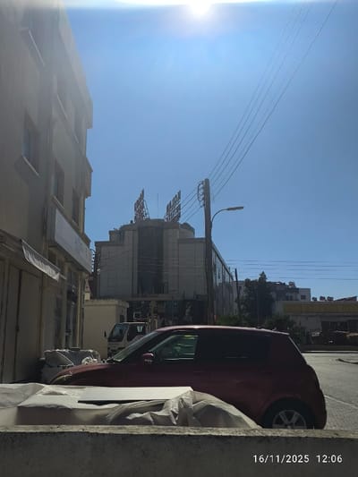 This image depicts an urban street scene in Gonyeli, Northern Cyprus / Cyprus, under bright, clear blue skies with a prominent sun flare in the upper center. The timestamp "16/11/2025 12:06" is visible in the lower right corner.

In the foreground, a low concrete barrier or wall is visible, with several white, wrapped objects or packages resting on or immediately in front of it. Directly behind this barrier, a dark red, two-door hatchback vehicle, possibly a Suzuki Swift, is parked or stationary, facing towards the right.

To the left, a multi-story building with a light-colored facade and visible windows occupies the edge of the frame. In the center-right, a dark utility pole supports multiple overhead power lines that extend diagonally upwards. A single street light fixture is attached to this pole.

The dominant structure in the midground is the Kolan British Hospital, identified by its modern architecture featuring a glass facade, tiered upper levels, and red illuminated "KOLAN HOSPITAL" signage on its roof. A white light commercial vehicle is partially visible near the base of the hospital building. Further in the background, other distant buildings and sparse green foliage are discernible. No individuals are visible within the frame.