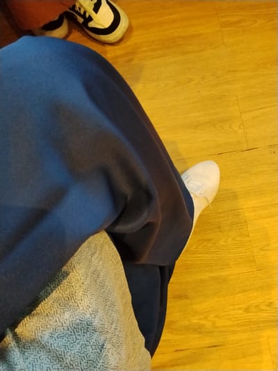 The image shows a person's legs and feet, dressed in dark blue pants and white sneakers. The person is seated on a textured light-colored cushion or blanket, with their legs extended onto a polished wooden floor. In the background, another pair of shoes, black and white sneakers, are visible. The lighting in the scene is warm and casts distinct shadows, suggesting an indoor environment. There is no visual information in the image that allows for the determination of the specific location within the city of Serang, Indonesia.