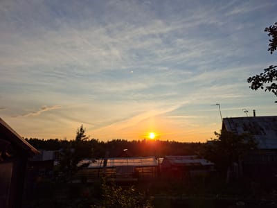 This image captures a serene sunset over a rural landscape in Pervouralsk, Russia. The sky is a gradient of soft blues and oranges, with wispy clouds streaking across the expanse. A faint moon is visible in the upper left quadrant of the sky, adding to the ethereal feel of the scene.

The foreground is dominated by the silhouettes of various structures, suggesting a residential or dacha area. To the left, the side of a dark wooden building is visible, its roofline extending into the frame. In the center and right, several greenhouses with translucent coverings are nestled amongst trees and other buildings. One building on the far right has a weathered, corrugated metal roof and a chimney, hinting at a permanent dwelling. An antenna is affixed to this roof, a common sight in rural settings.

The setting sun casts a warm, golden glow on the horizon, silhouetting a dense line of trees that stretches across the middle ground. The overall impression is one of tranquility and the peaceful end of a day. The light suggests it is late afternoon or early evening. No people are visible in the frame, reinforcing the sense of quiet solitude. The location context of Pervouralsk, Russia, places this scene in an area known for its natural beauty and outdoor living. The image evokes a feeling of calm and rustic simplicity. There is no visible text in the image.