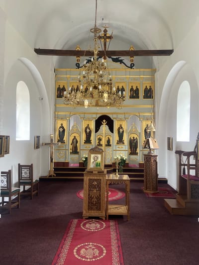 The image depicts the interior of an Orthodox church in Kolašin, Montenegro. The architecture features white-painted walls and arched ceilings. A prominent iconostasis adorned with gold accents and religious paintings dominates the foreground. A large, ornate chandelier hangs from the ceiling. Religious objects like a wooden lectern and a red carpet with golden decorations are visible, implying a place of worship. A wooden cross hangs against the wall. Natural light enters through arched windows. No people are visible.