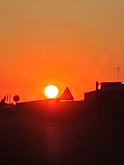 This is a photograph of a sunset in Valverde de Leganés, Spain. The sun is a bright white orb, low on the horizon, casting an intense orange and red glow across the sky. Silhouetted against the vibrant sky are several objects: a triangular traffic sign, the roofline of a building with an antenna, and some smaller structures that appear to be streetlights or poles with a red spherical object. The foreground is dark, obscuring details of the landscape or any people present. The overall impression is one of a warm, dusky evening in an urban or semi-urban environment.