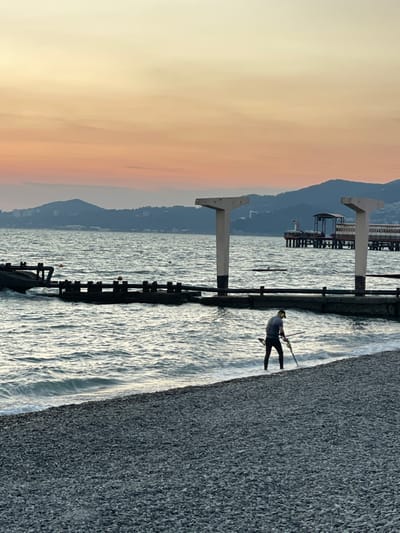 In the city of Progress, Russia, a lone individual stands on a pebble beach, wading into the gentle waves of the sea at dusk. The sky above is painted with soft hues of orange and yellow, transitioning into a pale cream towards the horizon. In the distance, a silhouette of mountains lines the background, with a pier extending out over the water. The pier features a structure with a roof and seating, and two large concrete pillars with crossbeams stand prominently in the mid-ground, one on each side of the frame. The person is wearing dark clothing and appears to be holding a long, slender object, possibly a pole or a tool, possibly engaged in an activity such as metal detecting or collecting objects from the water. The sea is calm with small waves lapping at the shore. The beach is covered in small, grey pebbles.
