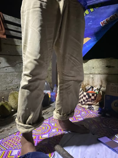 The image shows the lower legs and feet of an individual wearing light-colored, cuffed pants. They are standing on a patterned mat with purple, yellow, and white geometric designs. To the left, two durians are visible. In the background, there is a wooden wall, a blue tarp with lettering, a checkered shopping bag, and various bottles. The scene appears to be outdoors at night, possibly in Lamtui, Indonesia, given the presence of durians.