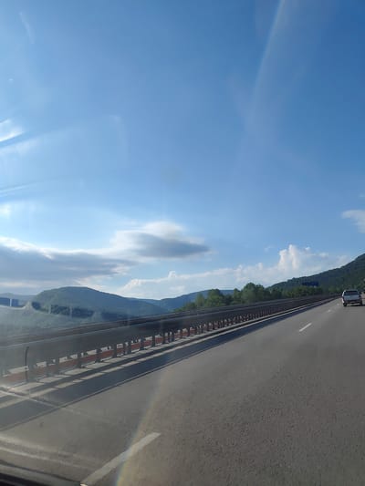 The image shows a view from a vehicle traveling on a highway. The highway has two lanes and a metal guardrail separating the lanes from the embankment. The embankment is covered with greenery and trees, with mountains in the background. The sky is blue with some clouds. A white SUV is visible on the highway ahead. The image is likely taken in Vrachesh, Bulgaria, based on the presence of a highway and mountainous terrain typical of the region.