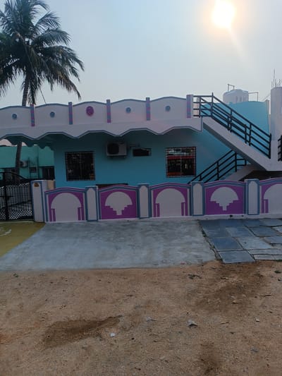 The image presents a brightly colored, two-story building located in Kothacheruvu, India. The building's main facade is painted in shades of light blue and white, adorned with decorative purple accents on the fence and roofline. Two barred windows are visible on the first floor. A black metal staircase leads to the second story, which also features a water tank on the roof. An air conditioning unit is mounted on the wall.
In the foreground, the ground is a mixture of concrete, stone tiles, and dirt. A tall palm tree is to the left of the building. The sky is clear with the sun positioned near the top of the frame. A neighboring building with similar architectural style and paint color is visible to the left of the main structure, partially obscured by a black metal gate.
