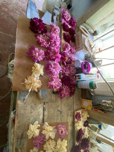The image shows a close-up, overhead view of a wooden workbench cluttered with various objects, including a collection of flowers, bottles, and tools. The workbench itself is old and weathered, with visible wear and tear. On the left side of the image, a large collection of pink and magenta flowers is arranged on a piece of brown cardboard. Some of these flowers appear fresh, while others are dried or wilting. To the right of the flowers, several plastic containers and bottles are scattered across the workbench. Among these are a can of spray paint, a tube of glue, and what appear to be small cups. Further to the right, a window with visible blinds is partially visible, allowing natural light to illuminate the scene. The overall impression is one of an artist's or crafter's workspace, possibly in the process of preparing or preserving flowers. There is no indication of specific people or actions, nor is there any discernible geographical context provided within the image itself. The location of Negrești, Moldova, is not evident from the visual information provided.