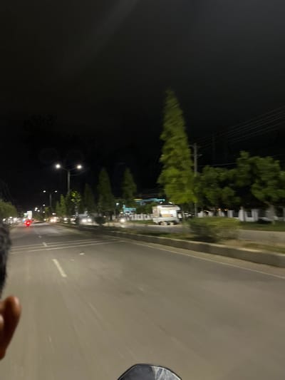 A first-person perspective shot from a moving vehicle at night in Tanjungkarang, Indonesia. The road is paved with lane markings and lined with trees and streetlights. In the distance, cars are visible. The overall lighting is dim, with streetlights and vehicle headlights illuminating the scene. The image has a slightly blurred quality due to motion.