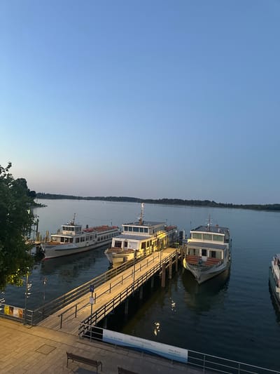 This is a photograph taken at dusk at the harbor of Prien am Chiemsee, Germany. The image shows several passenger boats docked at a wooden pier. The sky is a clear, pale blue, transitioning to a lighter hue near the horizon. The lake water is calm and reflects the lights from the pier and boats.
The main subjects are three passenger boats. The largest boat, labeled "BERTA," is positioned closest to the viewer and is white with a red stripe along its waterline. It has multiple decks and windows. Two other boats are visible, one partially obscured by the "BERTA" and another further down the pier to the right, which is also white. The wooden pier extends into the water and is illuminated by lights, casting reflections on the water. There are metal railings along the sides of the pier.
In the foreground, a paved walkway with benches can be seen. There are also signs on poles along the pier, one with the number "6" and another with "7". A banner with German text, including "Herrenchiemsee Festspiele" and "Der Welt entrückt", is attached to the railing of the pier.
On the left side of the image, there is a large tree with dark green foliage, partially obscuring the background. Across the lake, a treeline is visible in the distance, suggesting the shore of the lake. The overall atmosphere is serene and peaceful, indicative of the end of a day at a popular tourist destination. There are no visible people in the photograph.