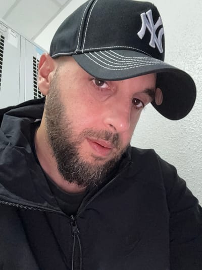 A man with a beard and a black baseball cap with the New York Yankees logo is shown from the chest up, facing the camera. He is wearing a black jacket with a zipper. The background appears to be an indoor setting with white textured walls and metal lockers. The lighting suggests an artificial light source. The scene is not identifiable as being in the city of Emmen, Netherlands.