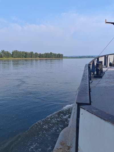 A wide, calm river flows under a blue sky with scattered white clouds. The river is bordered by a lush green treeline on the far bank. In the foreground, the side of a large vessel is visible, its grey hull extending into the water. Waves are created by the vessel's movement, seen as white foam churning beside it. The deck of the vessel features large, dark bollards. The scene depicts a journey on a river, likely in a natural or semi-rural setting, with no people or specific landmarks of a city visible.