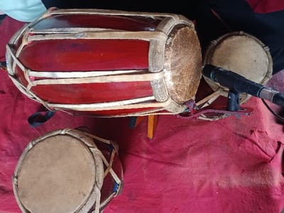 The scene in Kawungluwuk, Indonesia, displays three traditional Indonesian kendang drums arranged on a textured red fabric surface. The prominent object is a large, horizontally oriented kendang featuring a dark red lacquered wooden body with visible wood grain, secured by light tan rawhide lacing over tan drumheads. This instrument rests on a partial light brown wooden stand. To the lower left, a smaller, dark red kendang with similar rawhide lacing and drumheads is positioned vertically. A third, similarly constructed smaller kendang is partially visible behind the large instrument. A black dynamic microphone with an attached XLR cable is mounted on a small, black and red stand, positioned directly in front of the right drumhead of the large kendang, suggesting preparation for audio recording or amplification. The red fabric underlay exhibits folds and minor discoloration. No human subjects are present; the arrangement indicates a setup for a musical performance or recording.