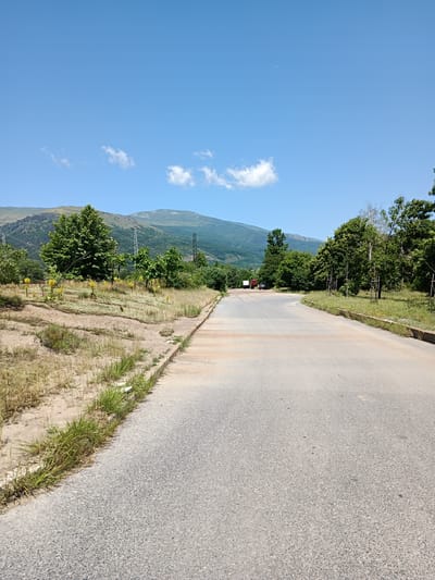 The image shows a paved road curving to the right, bordered by dry grass and dirt on the left and a grassy area with trees on the right. In the background, there are several trees, power lines, and mountains under a clear blue sky with scattered clouds. Two red trucks and a black car are visible in the distance on the road. This scene is located in the city of Zlatitsa, Bulgaria.