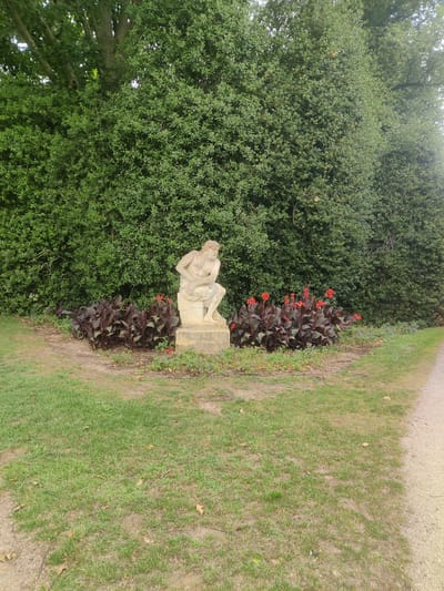 This image depicts a statue of a seated, contemplative male figure in a garden setting. The statue is made of light-colored stone and rests on a pedestal. Behind the statue, there is a dense hedge of green foliage, and in front of it, a cluster of plants with dark leaves and bright red flowers. The ground is covered in grass, with some patches of soil and scattered fallen leaves. To the right, a gravel path runs diagonally through the scene. The overall impression is one of a peaceful, manicured outdoor space. The location is identified as Isselburg, Germany, though no specific landmarks or street names are visible in the image. There are no people present in the image, and no actions or interactions are depicted.