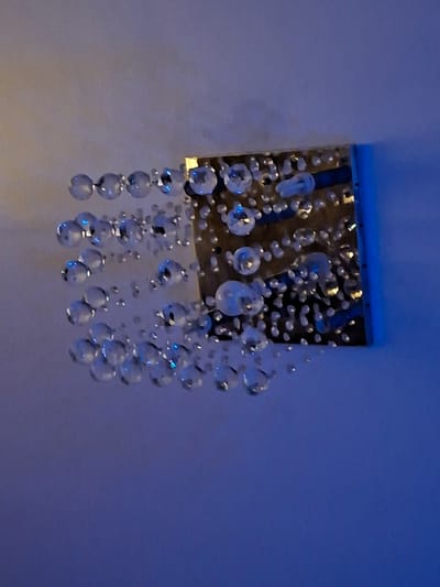 The image shows a close-up of a decorative wall fixture with numerous clear, spherical crystal beads hanging from a metallic rectangular base. The beads vary in size and are arranged in clusters and strands, catching and reflecting light, creating a sparkling effect. The background is a smooth, light-colored wall, with a gradient of blue light illuminating the scene from the right side of the image. The fixture appears to be mounted on a wall. The location is not specified in the image itself, therefore, a specific location in Evere, Belgium, cannot be provided.