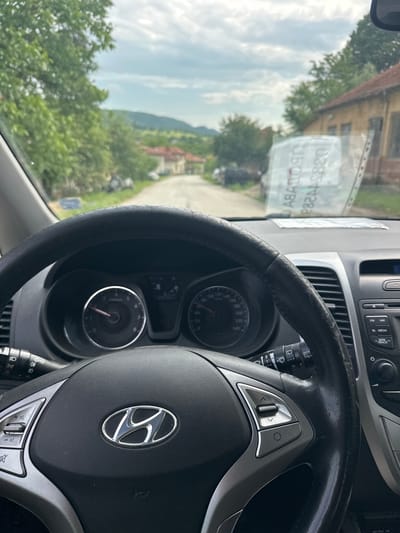 The image captures a view from inside a car, looking out through the windshield. In the foreground, a black steering wheel with a silver Hyundai logo at its center dominates the right side. The steering wheel appears somewhat worn, and various control buttons, including those for cruise control, are visible on its right spoke. To the left of the steering wheel, a wiper stalk is visible.
Behind the steering wheel, parts of the car's dashboard are visible, including the speedometer, which indicates a maximum speed of 240 km/h, and a tachometer. Above the steering wheel, part of the infotainment system is seen, with buttons for "FM," "AM," "MEDIA," and "SEEK TRACK."
Through the windshield, a bright daytime scene unfolds. A road can be seen in the mid-ground, leading into a rural or semi-rural landscape. Lush green trees and vegetation are prominent, especially on the left and center. In the distance, hills or mountains rise under a cloudy sky. Several buildings with reddish roofs are visible among the trees, suggesting a village or small town setting.
Notably, mirrored text is reflected on the windshield. The visible text includes "ABATON" and the number "03851283," which resembles a phone number. These reflections suggest a sticker or sign placed inside the car, facing outwards. The overall impression is that of being parked or driving slowly in Dobrodan, Bulgaria, offering a glimpse of the local scenery.