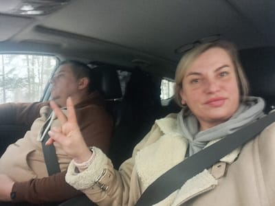Two people are inside a car. The driver, a man, is focused on the road. The passenger, a woman, is turned towards the camera, making a peace sign. Both occupants are wearing seatbelts. The woman has blonde hair, a fair complexion, and is wearing a beige jacket and grey hoodie. The man is wearing a brown hooded sweatshirt. Through the car windows, blurry trees are visible. The interior of the car is grey and dark.