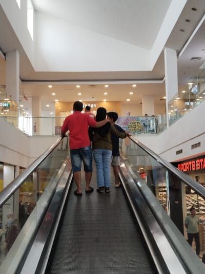 The image captures a perspective from an escalator in a mall in Kudus, Indonesia. The escalator is moving upwards, with a group of three people in the foreground, viewed from behind. A man in a red shirt and denim shorts is on the left, with his arm around a woman in a green shirt and jeans. To their right, a young person in a dark blue shirt and grey shorts is also on the escalator. In the background, the interior of the mall is visible, with shops on multiple levels. A sign for "SPORTS STATION" is prominent on the right. The overall environment suggests a typical shopping mall setting with overhead lighting and architectural features.
