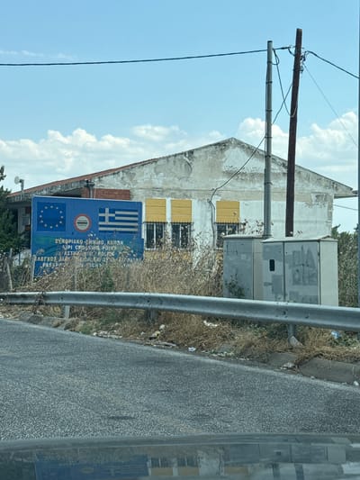 This is a medium shot, outdoor, eye-level, photographic image of a border crossing in Kipi, Greece. The scene is dominated by a blue sign with the European Union and Greek flags, indicating a border control point. Text on the sign is visible in Greek and English, reading "ΕΛΕΓΧΟΣ CONTROL" and "ΣΥΝΟΡΙΑΚΟ ΣΗΜΕΙΟ ΕΛΕΓΧΟ Kipi Crossing Point". The building behind the sign is weathered and appears to be a derelict structure with faded paint and yellow shutters. Two gray electrical boxes are positioned in front of the building. A metal guardrail runs across the foreground, separating the road from the area with the sign and building. The environment is characterized by dry, overgrown vegetation and scattered litter, suggesting a somewhat neglected area. The sky is partly cloudy. There are no people visible in the frame.