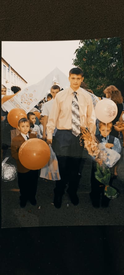 The photograph depicts a group of people, likely students and perhaps teachers, gathered outdoors. In the foreground, a young boy in a brown blazer and tie holds a large, orange balloon. To his right, a taller young man in a light peach-colored button-down shirt and a patterned tie stands prominently. He is holding a bouquet of flowers. Behind them, other children and adults are visible, some holding balloons, including a pale pink one with a cartoon image. A white banner or cloth with an embroidered pattern is held aloft by some individuals in the background, suggesting a celebratory or ceremonial event. The setting appears to be a street or walkway in front of a building with windows, possibly a school. Trees and foliage are visible in the background, and the lighting suggests it is daytime, likely with overcast or diffused light. The overall atmosphere seems to be one of a school event, possibly the start of the school year given the presence of balloons and flowers. There is no visible text in the image.