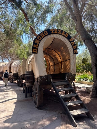 The image shows covered wagons with "Rancho Texas Park" signage. The location is likely Rancho Texas Lanzarote Park in Tías, Spain. The wagons are in an outdoor area with trees and paved ground. There is a person in the lower section of the photograph.