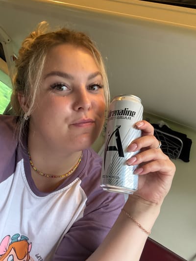 In the city of Doma 1079 Km, Russia, a young Caucasian woman with blonde hair styled in a bun is shown in a selfie. She is wearing a purple and white raglan t-shirt with a cartoon character graphic. Around her neck, she wears a colorful beaded necklace, and on her left wrist, a delicate rose gold bracelet. Her right hand is holding a silver can of "Adrenaline Zero Sugar" energy drink. The background appears to be the interior of a train carriage, with visible seating and overhead compartments. The lighting suggests it is daytime.