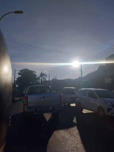 The image depicts a bright, sunny day with a low sun angle, suggesting either early morning or late afternoon. The scene is a street in Macege, Indonesia, with vehicles parked or driving along the road. Two cars are visible, one white and one silver pickup truck. The silver pickup truck has a license plate that reads "DD 8274 XZ". Overhead power lines are strung across the sky, common in many urban and semi-urban settings. Lush green trees, including a palm tree, are present, indicating a tropical or subtropical climate. The sunlight is intense and creates strong shadows, reflecting off the road surface. A street lamp is visible on the left side of the frame. The overall impression is of a typical day in a populated area with traffic and infrastructure.