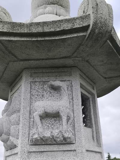 The image shows a close-up, low-angle view of a carved stone structure, likely a monument or part of a building, set against a cloudy sky. The stone is grey and appears textured, possibly granite. Prominently featured is a relief carving of a deer, standing with its head lowered, on one of the octagonal faces of the structure. To the right of the deer carving, there is an opening with a lattice-like pattern. The structure has multiple levels and architectural details, including a curved, tiered roof-like element at the top, with decorative carvings. The style suggests an East Asian influence. There are no people or animals present other than the carved deer. The environment appears to be outdoors, possibly in a park or garden, with a hint of greenery visible in the lower right corner, suggesting trees. The location is stated to be in Mažučiai, Lithuania, though the visual elements do not inherently confirm this geographical context.