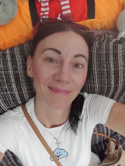 This is a selfie taken indoors, on a bed with a patterned throw. The subject is a woman lying down, looking directly at the camera. She has dark hair, brown eyes, and is wearing a white t-shirt. A thin strap of a bag is visible across her chest, and she is wearing a necklace with a cross and a cloud pendant. Her face is lightly made up. The background includes colorful pillows and blankets. The lighting is bright and appears to be natural light. There is no discernible indication of the location being Priego De Córdoba, Spain.