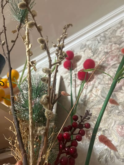 The image displays a close-up, upward-angled view of a static artificial floral and botanical arrangement situated indoors in Dmitriyevskaya, Russia. In the immediate foreground and mid-ground, numerous faux plant elements are visible: several bare branches adorned with light brown, fuzzy catkin-like structures; clusters of bright red, spherical artificial berries; sprigs of artificial green pine needles partially dusted with white material resembling snow; and various thin, dried-looking brown plant stalks and wispy, light orange or pink decorative grasses. Further back, out of sharper focus, are multiple bright red fuzzy spheres (possibly pom-poms) on slender green stems. On the far left, a dark blue or green object is partially visible alongside several bright yellow spherical decorations. The background features light-toned wallpaper with a subtle, embossed pattern, likely floral or damask, in muted pink, beige, and gold hues. A white, decorative crown molding runs horizontally along the top of the wall, separating it from the ceiling. No individuals are present in the scene, and no discernible actions or interactions are occurring.