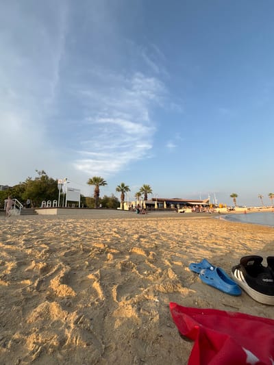 This is a full-frame, eye-level shot of a sandy beach in Toulon, France, with the water and a restaurant in the background. The foreground is dominated by sand, with a red towel and a pair of blue clogs and black Adidas sneakers placed on it. In the midground, a raised concrete walkway separates the sand from a grassy area lined with palm trees. Several people are visible on the walkway and in the grassy area, some sitting and some walking. A white sign with a red symbol is mounted on a pole. Beyond the walkway and trees, a long, low building with a brown roof, presumably a restaurant, is visible, with yellow umbrellas and people seated at tables. Further in the background, the Mediterranean Sea is visible with a few boats and people in the water. The sky is a clear blue with some scattered white clouds. The lighting suggests it is late afternoon.