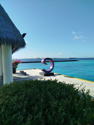 This is a photograph of a waterfront scene in Gulhi Falhu, Maldives. In the foreground, a dense, dark green hedge runs horizontally across the bottom of the image. Behind the hedge is a wooden pier, composed of light-colored planks with a yellow painted edge. On the pier, a large, purple, spiral sculpture is positioned on a dark rectangular base. To the left of the sculpture, and partially obscured by it, are a few dark gray steps. To the far left of the image, a thatched roof structure with a white support pillar is visible, with a black decorative element on the roofline. A large dark pot sits at the base of the pillar, filled with vibrant pink and white flowers. Beyond the pier and sculpture, the turquoise waters of the ocean stretch to the horizon. A dark, thin breakwater runs across the middle ground of the water, with several small structures visible on it. In the far distance, a hazy cityscape with buildings and industrial structures can be seen on the horizon. The sky is a clear, bright blue with a few wispy white clouds. The lighting suggests it is daytime.