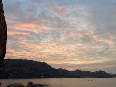 The scene, situated in Hale Bandi Harlapura, India, captures a natural landscape during twilight, characterized by a low sun angle. The lower half of the frame is occupied by a calm body of water. The sky above features a dense formation of clouds displaying shades of pink, orange, and light grey against a pale blue background. A dark, vertical rock face is partially visible along the far left edge of the image. Numerous large, smooth boulders are positioned at the water's edge and partially submerged in the foreground. Across the water, a series of dark, rugged hills, sparsely covered with vegetation, forms the horizon. No individuals, human activity, or man-made objects are present within the frame.