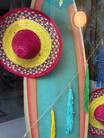 The image shows a close-up of a decorative display, likely in a shop window, featuring Mexican-themed items. Dominating the frame is a large, colorful sombrero with concentric circles of yellow and pink straw, edged with a diamond pattern of pink and green. Below it, part of another similar sombrero is visible on the right. Hanging on thin strings are several bright, textured tassels in shades of yellow, turquoise, and green. In the background, a surfboard-like object with rainbow stripes in pink, orange, yellow, and green stands vertically. Behind the surfboard, a reflection shows palm trees and what appears to be a circular light fixture. The overall impression is festive and summery. The location is a shop window in Strombeek-Bever, Belgium.