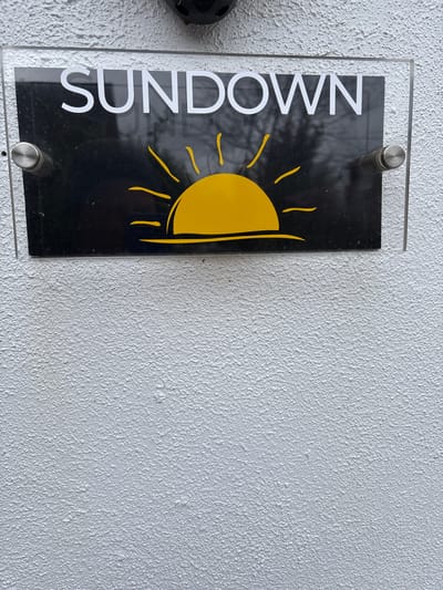 A sign reading "SUNDOWN" with a yellow sun graphic is mounted on a white, textured wall. The sign is black with white lettering, enclosed in a clear acrylic frame affixed to the wall with metal standoffs. Part of an unidentifiable black object is visible on the left edge of the frame. The background of the sign includes reflections of trees. The photograph was taken in Longden, United Kingdom.