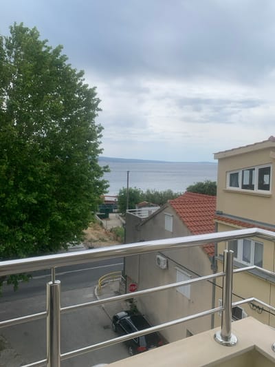 The image shows a view from a balcony in Dugi Rat, Croatia. The balcony has a stainless steel railing. The view looks out onto a street and a small neighborhood. In the distance, the Adriatic Sea can be seen. There is a large green tree in the foreground, a black car parked on the street, a "STOP" sign on the street, a light pole, and a house with an orange tiled roof. The sky is overcast and gray, with a hint of blue. There is a second house that has a yellow facade and a window with a wooden frame and glass panes. The house also has a large awning that is white. There are no people visible in the image.
