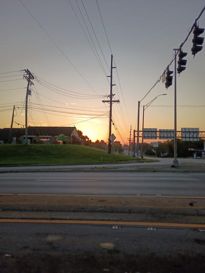 This media file captures a sunset scene in Frankfort, United States of America. The image is oriented vertically, with the top of the frame showing a clear sky transitioning to warm hues of orange and yellow as the sun sets. Several traffic lights are visible in the upper left, silhouetted against the bright sky. Numerous telephone poles and power lines crisscross the frame, indicating an urban or suburban setting.
In the mid-ground, a building with a sign that reads "WALK-IN DENTAL" is partially visible, surrounded by a grassy area. The road, with visible yellow lane markings, runs along the right side of the frame, appearing wet, suggesting recent rain or dew. The overall mood is calm and serene, enhanced by the soft lighting of the sunset. The time of day is clearly evening, and the weather appears clear, though the wet road implies some moisture in the air. The presence of traffic lights and the road suggests a public thoroughfare.