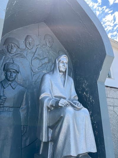 This media file is a close-up, angled shot of a relief sculpture. The sculpture is made of a dark material, possibly metal, and is painted in a reflective silver or gray color. The scene depicts several figures. In the foreground, a central figure is lying down, draped in what appears to be a cloak or uniform. This figure's head is slightly raised, and their eyes are open, looking forward. Their hands are clasped together in their lap, holding something indistinct. Behind this central figure, a row of faces emerges from the background. These are smaller, sculpted heads of individuals wearing what look like military helmets. Their expressions are somber or stoic. Below these, more figures are visible, also seemingly in military attire, with one figure's head prominently displayed, wearing a helmet with a star emblem. Some of these figures appear to be lying down or in a state of repose, with one holding what could be a rifle with a bayonet.

The overall mood conveyed by the sculpture is one of solemnity and remembrance, likely a memorial. The lighting suggests it is daytime, with bright sunlight reflecting off the sculpture's surface. The background shows a glimpse of a building and a blue sky with scattered white clouds, indicating an outdoor setting. The setting is identified as Takhtamukai, Russia. There is no visible text in the frame.