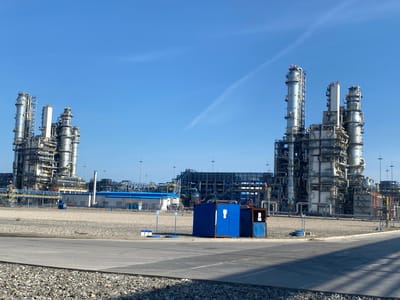The image depicts an industrial complex under a clear blue sky in Yukhta, Russia. In the foreground, a paved road separates the viewer from a landscape of gravel and a small blue utility structure with a dark blue door and a dark blue cover. Beyond this, a sprawling industrial facility dominates the scene. Two large, multi-story structures with numerous pipes, platforms, and vertical towers rise prominently. These structures are interconnected and feature a complex network of walkways and equipment. Further in the background, additional industrial buildings and more towers are visible, all part of the same industrial site. The overall environment is arid, with sparse vegetation, and the sky is a vibrant blue with a faint contrail. There are no visible people in the image.
