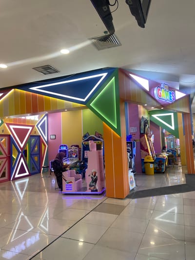 The image depicts the interior of an arcade, likely located in Kudus, Indonesia, indicated by the presence of the "MR. GAMES" sign and the overall aesthetic often found in such establishments in the region. The arcade features a colorful and modern design with geometric patterns and vibrant lighting. Arcade machines are visible, some displaying colorful graphics, and include a pink racing-themed game in the foreground. A woman wearing a hijab is interacting with one of the arcade machines. The floor is polished and reflects the lighting and triangular motifs. The environment appears clean and well-lit, typical of an indoor entertainment venue.