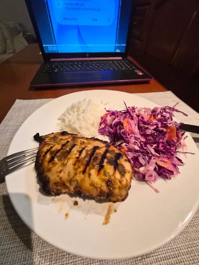 The image displays an indoor scene featuring a prepared meal and an active laptop on a wooden table. The specific location within the Municipal Unit of Corinth, Greece, cannot be determined from the visual evidence.

In the foreground, a white, round plate rests on a grey, textured placemat. The plate contains a grilled chicken breast, exhibiting distinct char marks and visible seasoning, alongside a portion of white rice. Adjacent to these, a salad composed primarily of shredded purple cabbage and orange slivers, likely carrots, is present. A silver four-tine fork is positioned on the left side of the plate, partially underneath the chicken. Minor food residue is visible on the plate beneath the chicken.

In the midground, a black HP laptop with a pink or purple lower chassis trim is open on a dark brown, polished wooden surface. The laptop screen emits a bright blue light and displays a user interface, consistent with a messaging application, showing fragmented text that includes "is Emily with you?" and "It's a surprise, so don't let her see!".

The background, largely out of focus, reveals a light-colored upholstered object on the left, possibly a chair or sofa, and darker wooden furniture or paneling on the right. No individuals are visible in the scene.