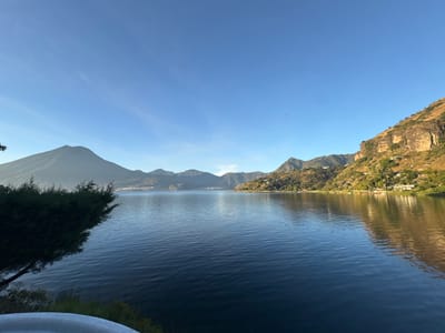 The image displays a panoramic landscape captured from San Pablo La Laguna, Guatemala, overlooking Lake Atitlán. The scene is dominated by the expansive dark blue waters of the lake, which occupy the central and lower portions of the frame, exhibiting subtle surface ripples. The sky above is clear and light blue, with minimal high-altitude cloud formations.

To the far left, a prominent, conical volcano, consistent with Volcán San Pedro, rises distinctly from the horizon, partially obscured by a slight atmospheric haze. Adjacent to this, other mountain ranges and distant lakeside settlements are discernible along the shore. The right side of the image features a steep, rugged hillside ascending directly from the lake's edge. This hillside is characterized by exposed, light-colored rock formations and interspersed green vegetation, illuminated by direct sunlight. Numerous small, light-colored buildings with distinct roofs are visible, nestled into the contours of this populated hillside.

In the immediate foreground, to the left, a dense mass of dark green evergreen foliage partially obstructs the view of the lake. At the very bottom edge of the frame, a curved, light-colored object, possibly a railing or part of a structure, is partially visible. No individuals or explicit actions are observed within the scene.