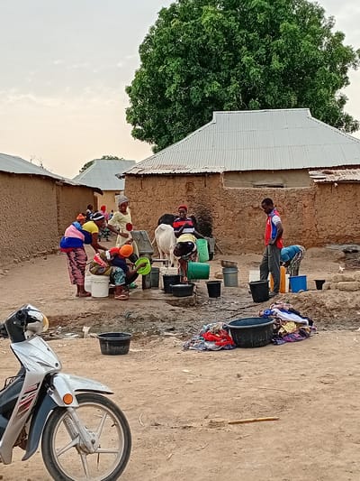 In Nata-Ala, Nigeria, a row of at least seven people, mostly women, are washing clothes outdoors. They are clustered along the side of a mud-brick building with a corrugated metal roof, each individual positioned in front of a bucket of water. The buckets are of varying colors and sizes, predominantly black, green, and blue. Clothes are piled on the ground beside the buckets. A man stands at the end of the row, also washing clothes. A motorcycle is parked in the foreground, partially obscuring the scene. A large tree is visible to the left of the building, and the sky is overcast. The ground is bare earth.